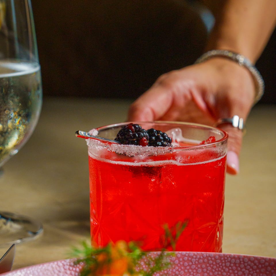 A vibrant red cocktail with blackberries and a sugar rim, held by a person's hand.
