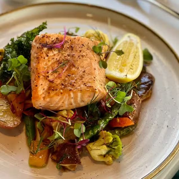 Pan-seared salmon with roasted vegetables, microgreens, and a lemon wedge on a white plate.