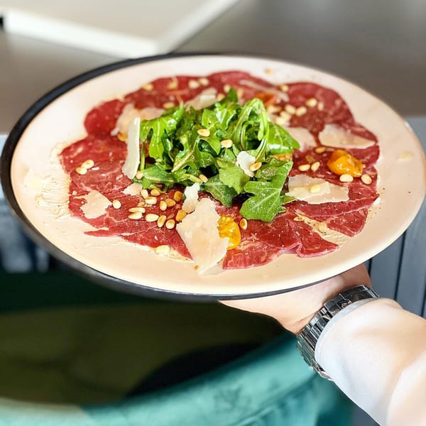 A hand holding a plate of Waxin's beef carpaccio with arugula, pine nuts, and parmesan.