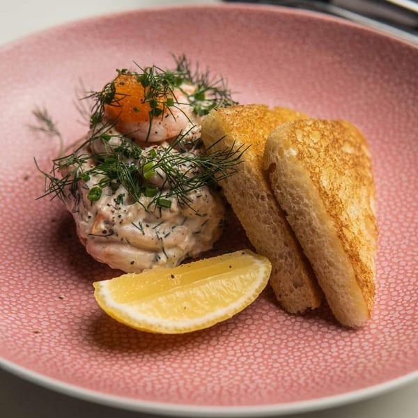 Shrimp skagen with dill, chives, and roe, served with toasted bread and a lemon wedge on a pink plate.