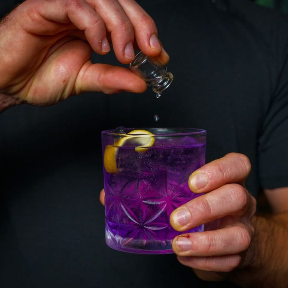 Hands pouring liquid from a small glass into a purple cocktail with lemon peel.