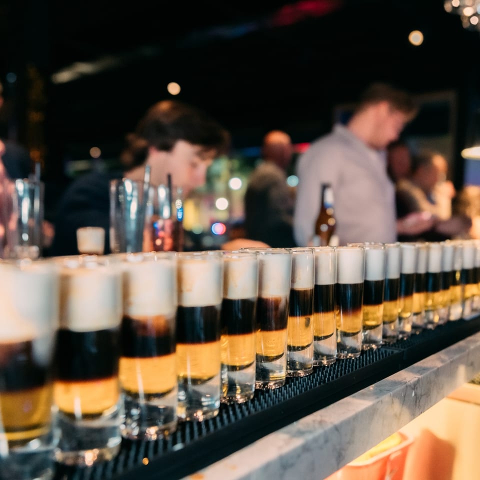 A row of layered shot glasses on a bar, with blurred patrons in the background at Waxin's.