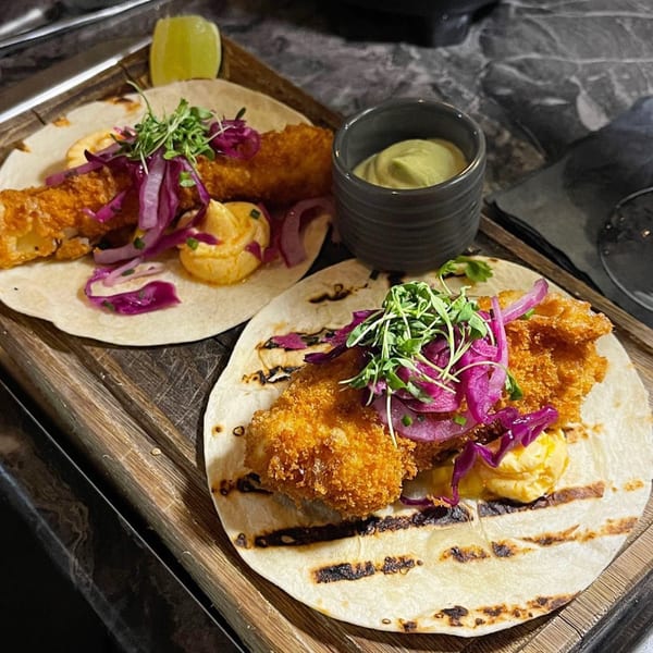 Two Waxin's fish tacos with fried fish, red cabbage, and sauce, served with a side of green dip.