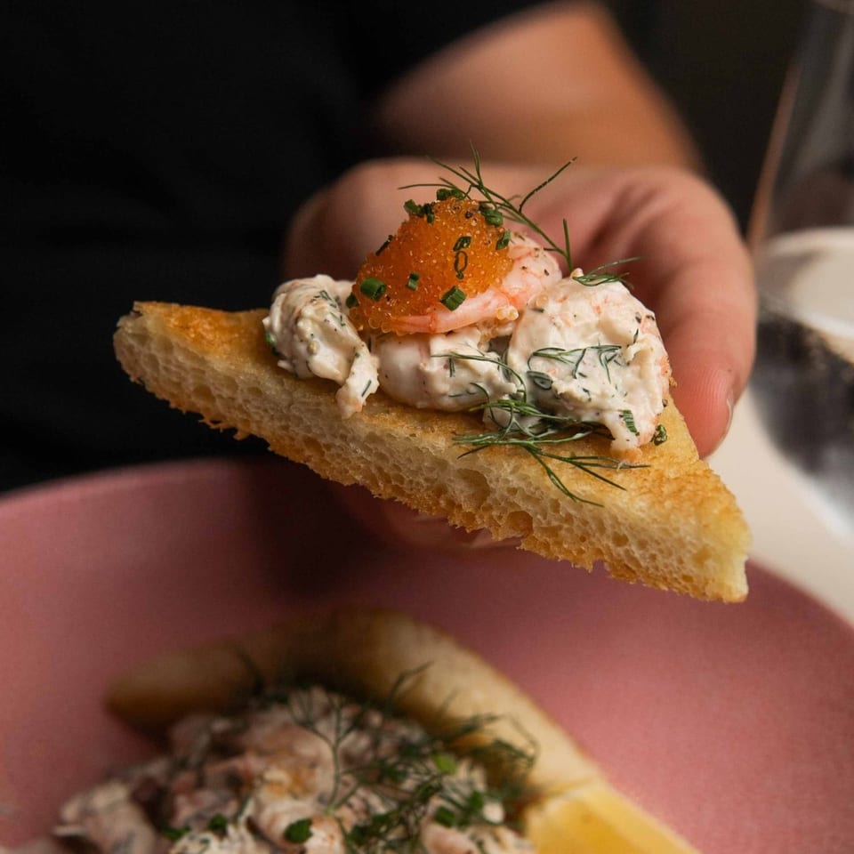 Hand holding a toasted bread point topped with shrimp, dill, and bright orange roe.