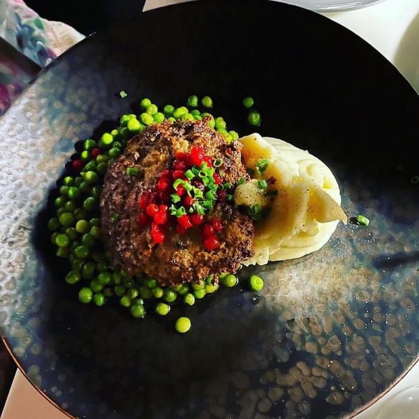 Meatloaf with red berries, chives, mashed potatoes, and green peas on a dark plate.